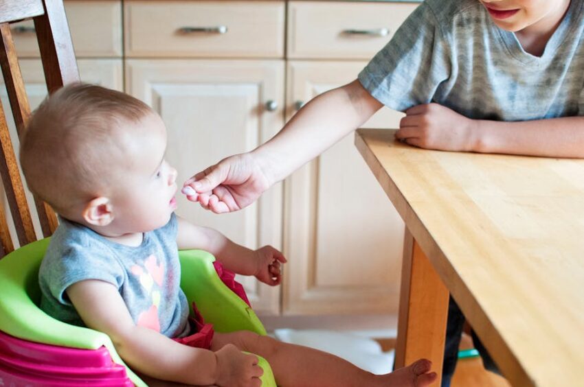 A cute baby in a high chair being fed by an older child in a bright kitchen setting.